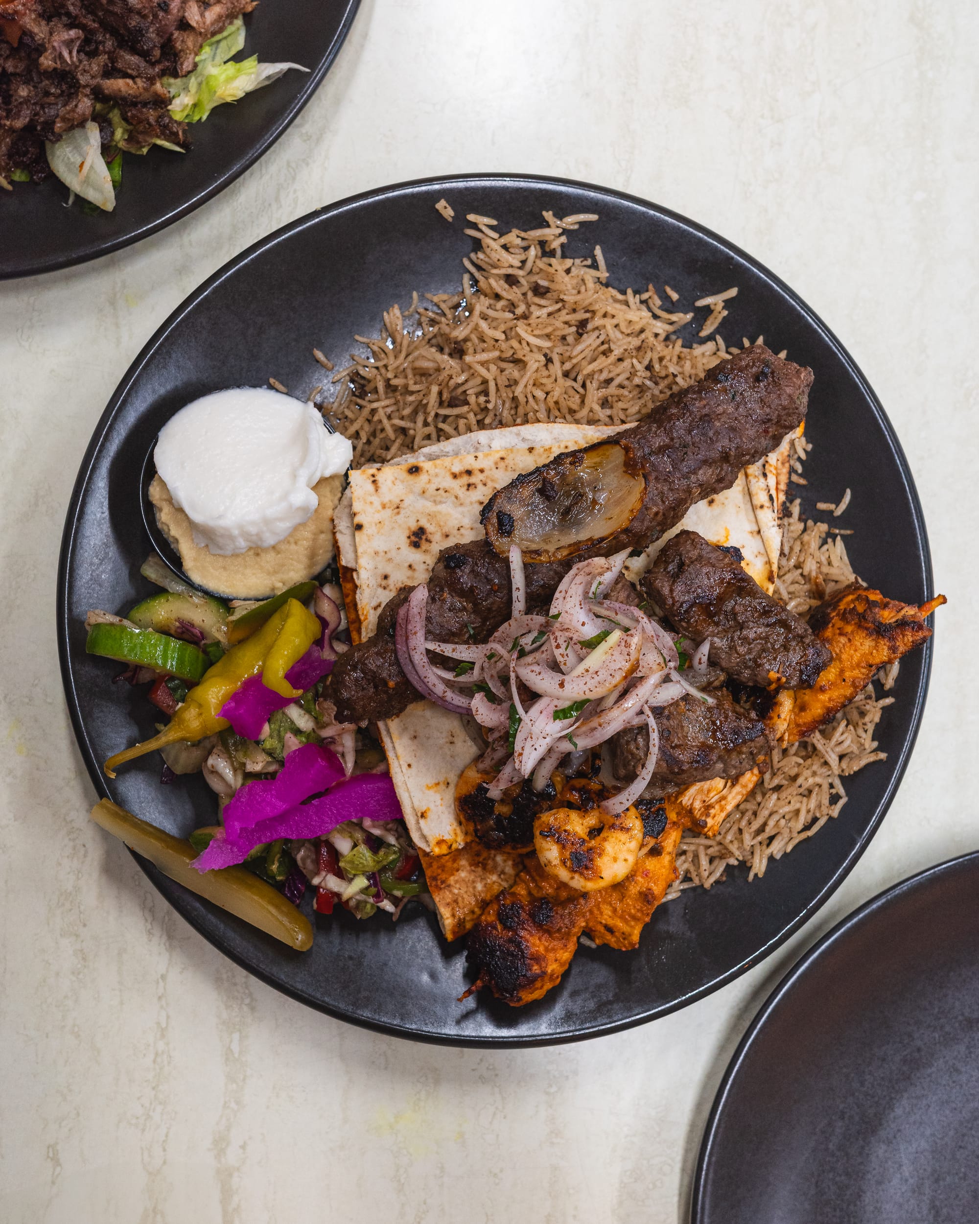 A plate of mixed grilled meats served with rice, pickled vegetables, and a side of hummus and onion dip.