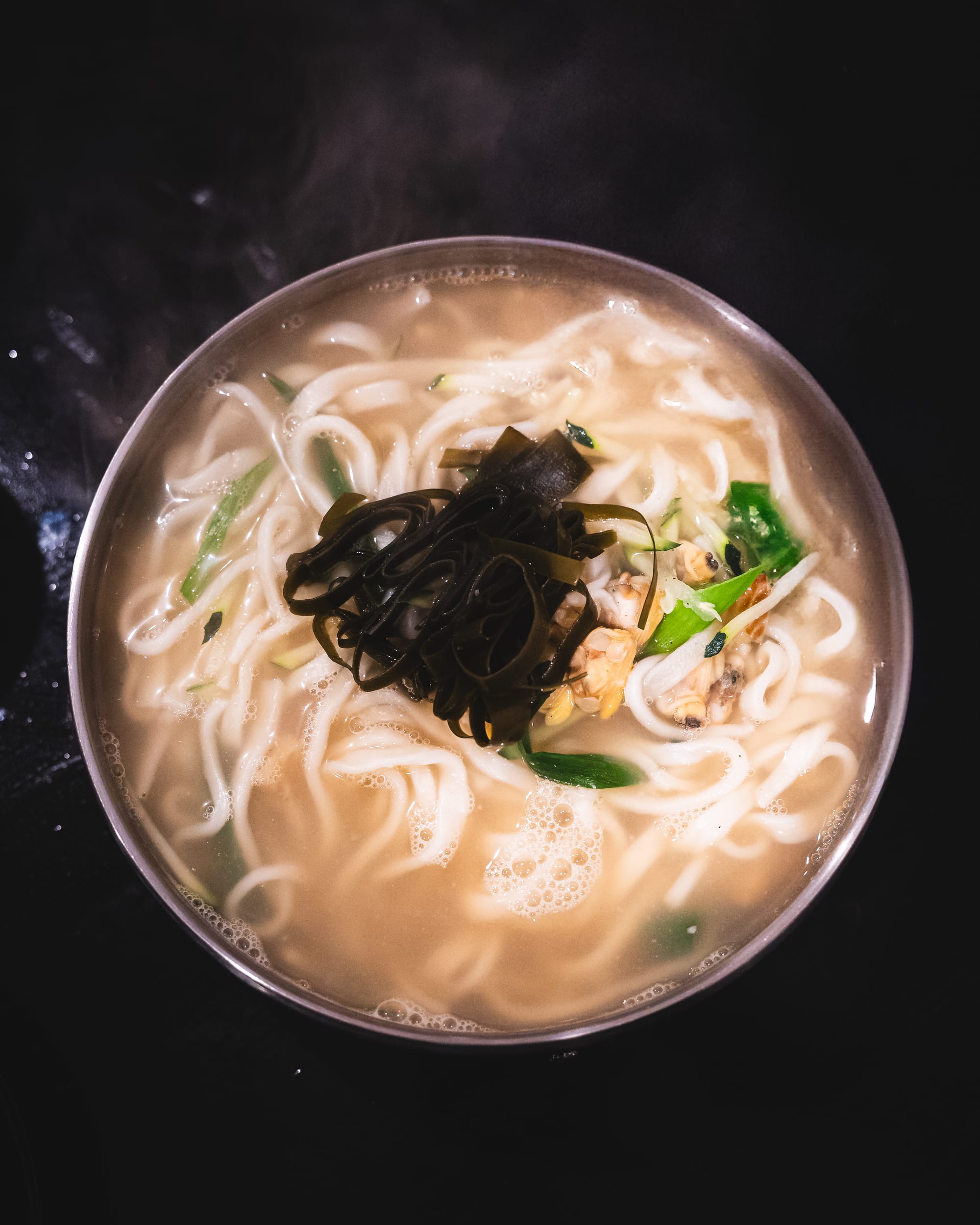 A bowl of steaming ramen with thin noodles, a clear broth, and green garnish.