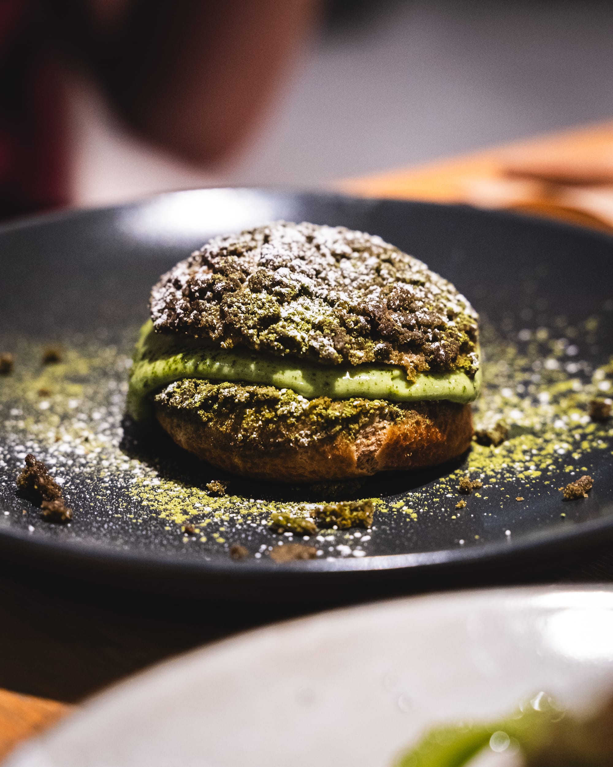 A matcha-infused cream puff dusted with powdered sugar, served on a black plate.