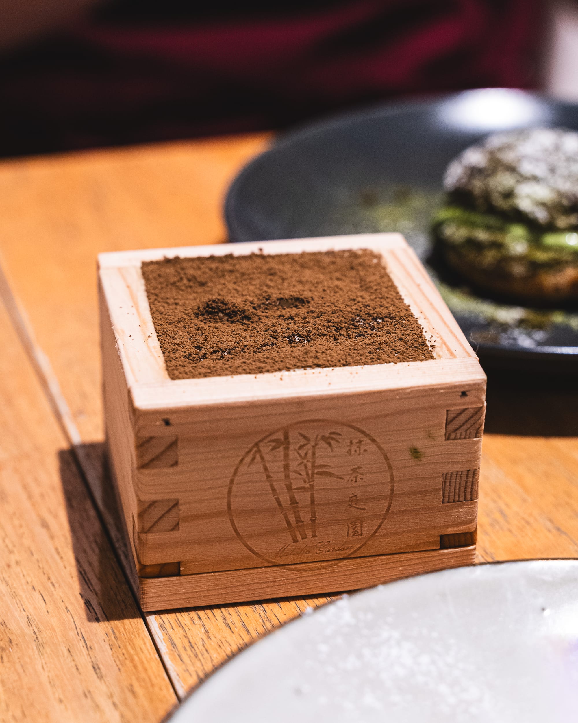 A wooden masu box filled with a finely dusted matcha dessert, placed on a restaurant table.