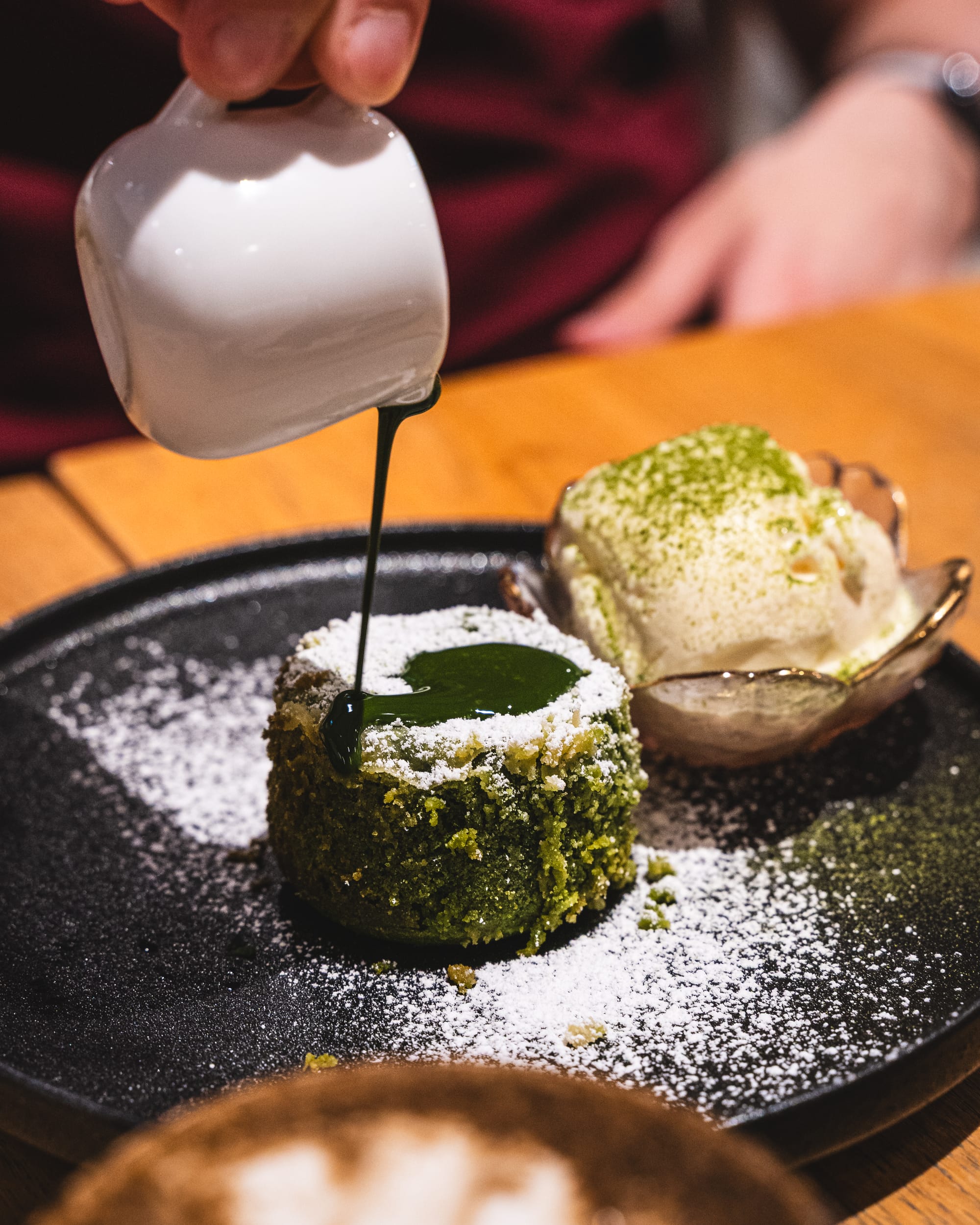 A hand pouring a sauce over a matcha-flavored dessert, garnished with crushed nuts and powdered sugar.