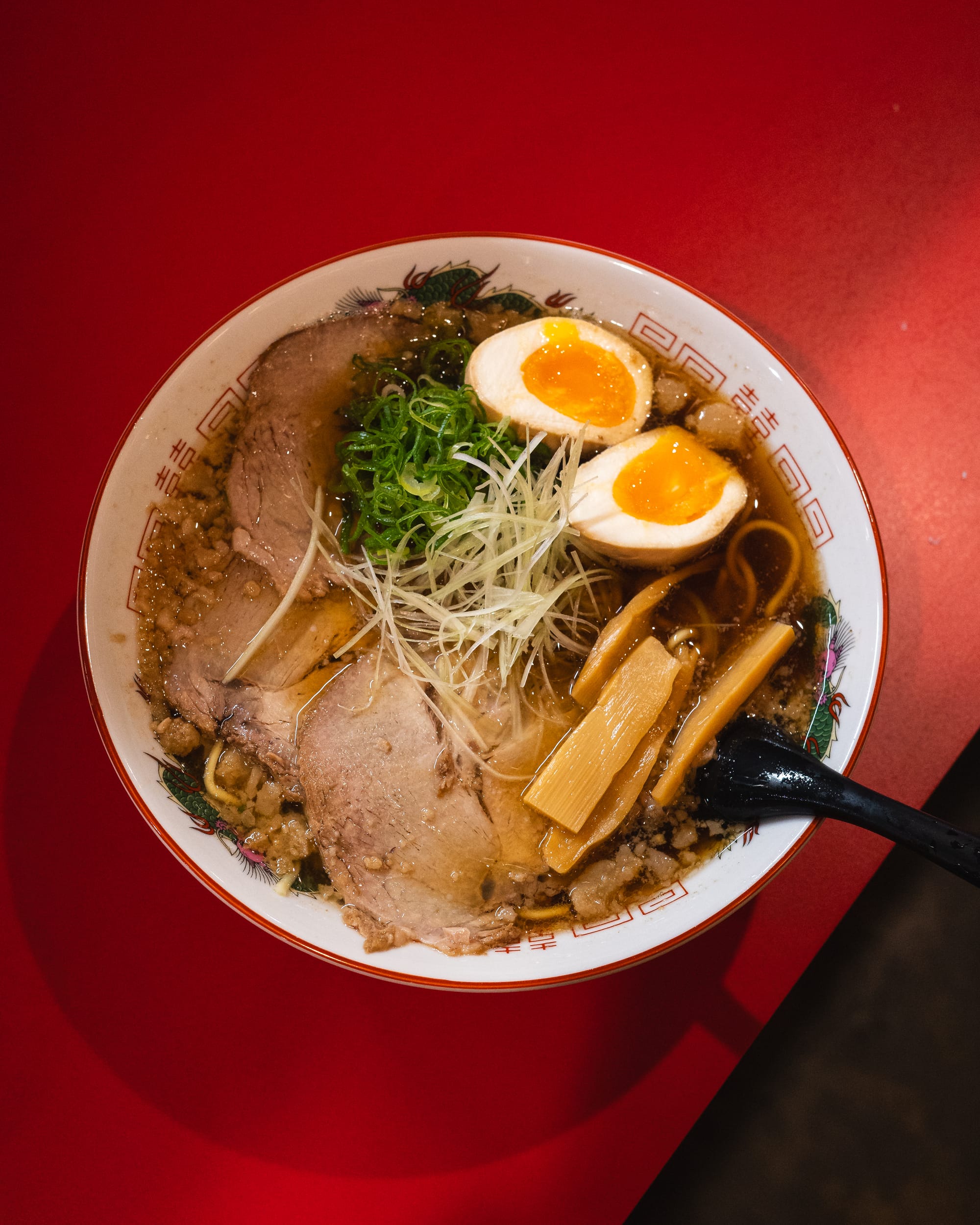 A bowl of ramen with sliced chashu pork, soft-boiled egg, bamboo shoots, green onions, and broth, served on a red table.