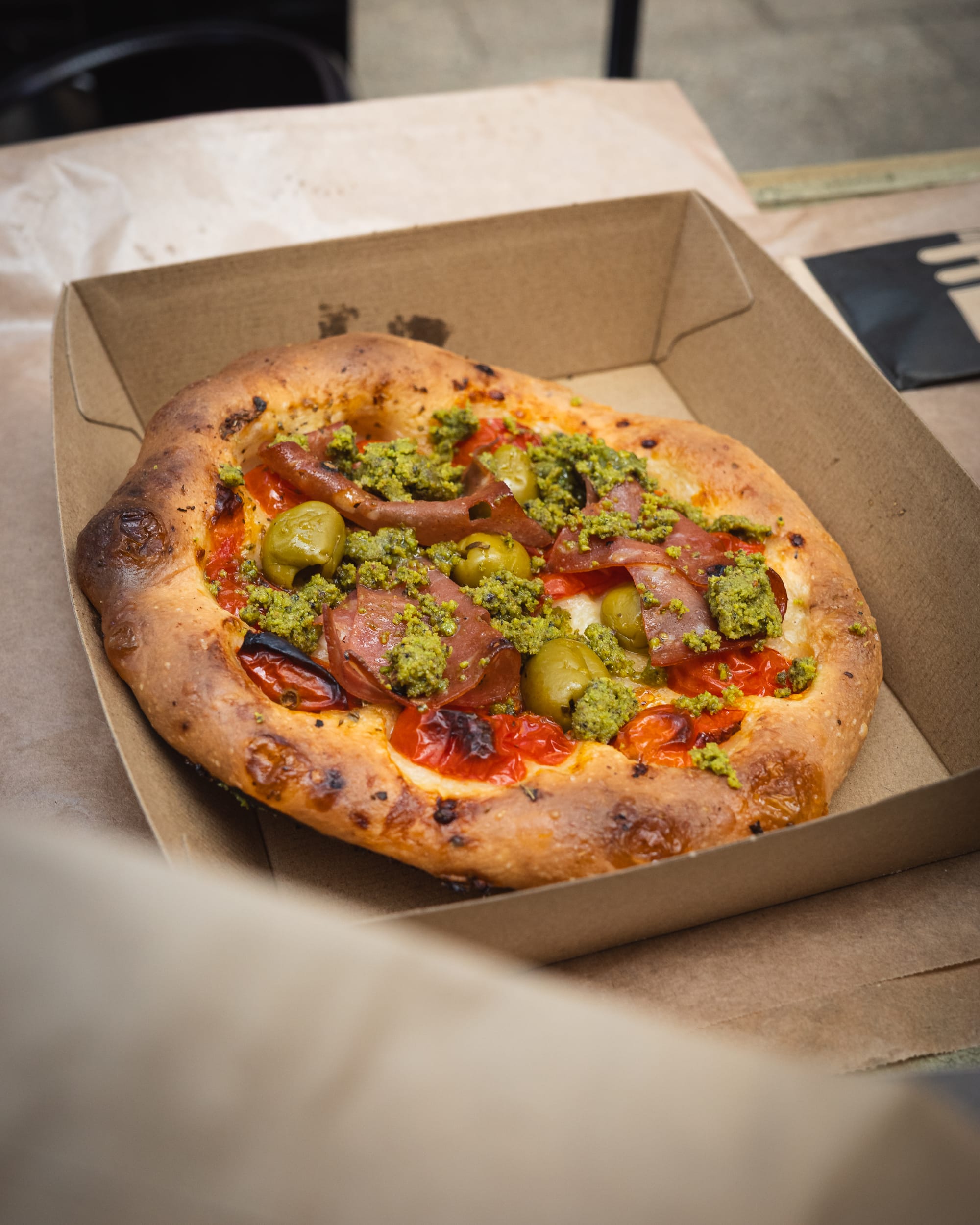 A small wood-fired pizza topped with cherry tomatoes, jalapeños, and melted cheese in a takeaway box.