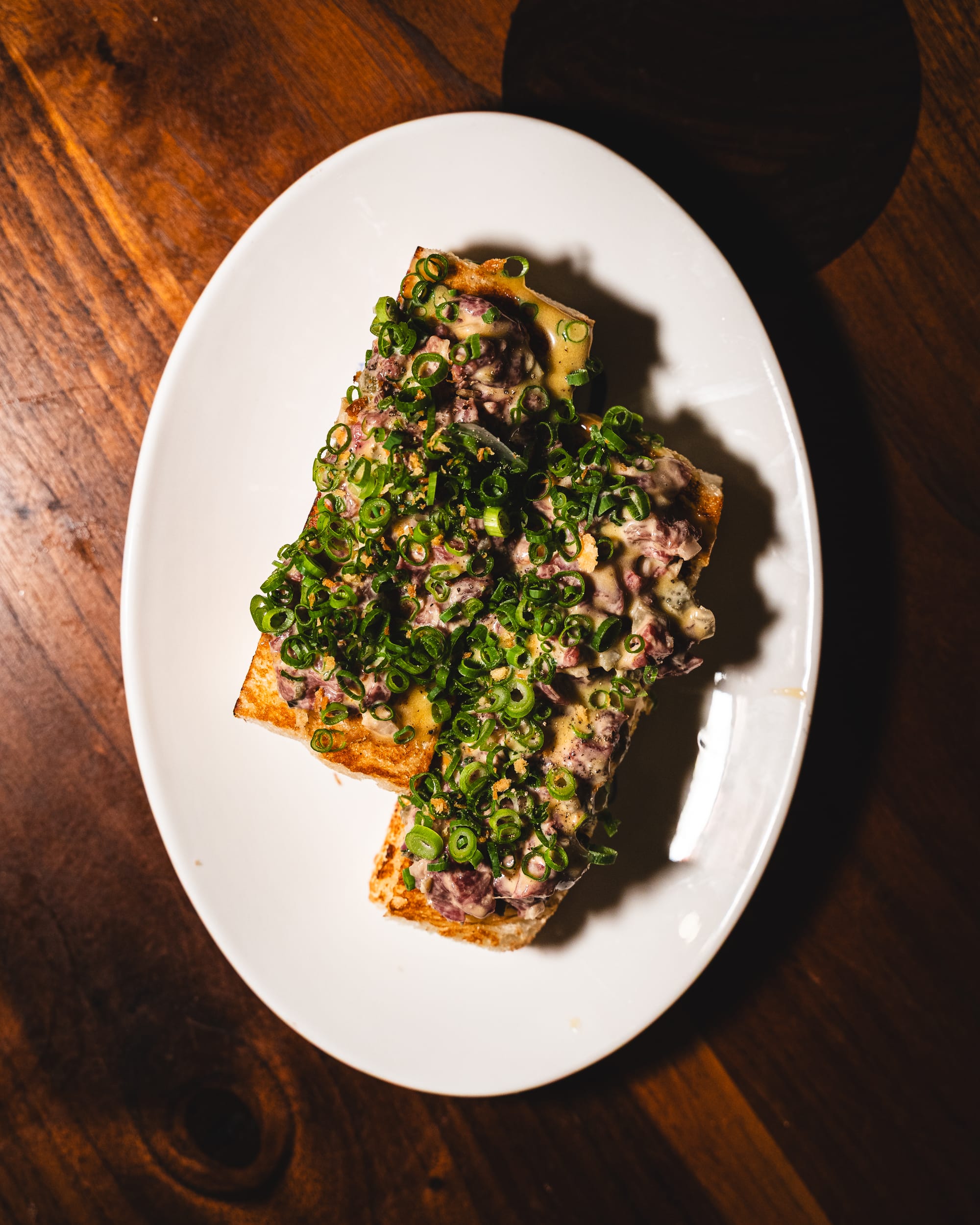 A plate of thick toast topped with green onions and beef tartare, served on a wooden table.