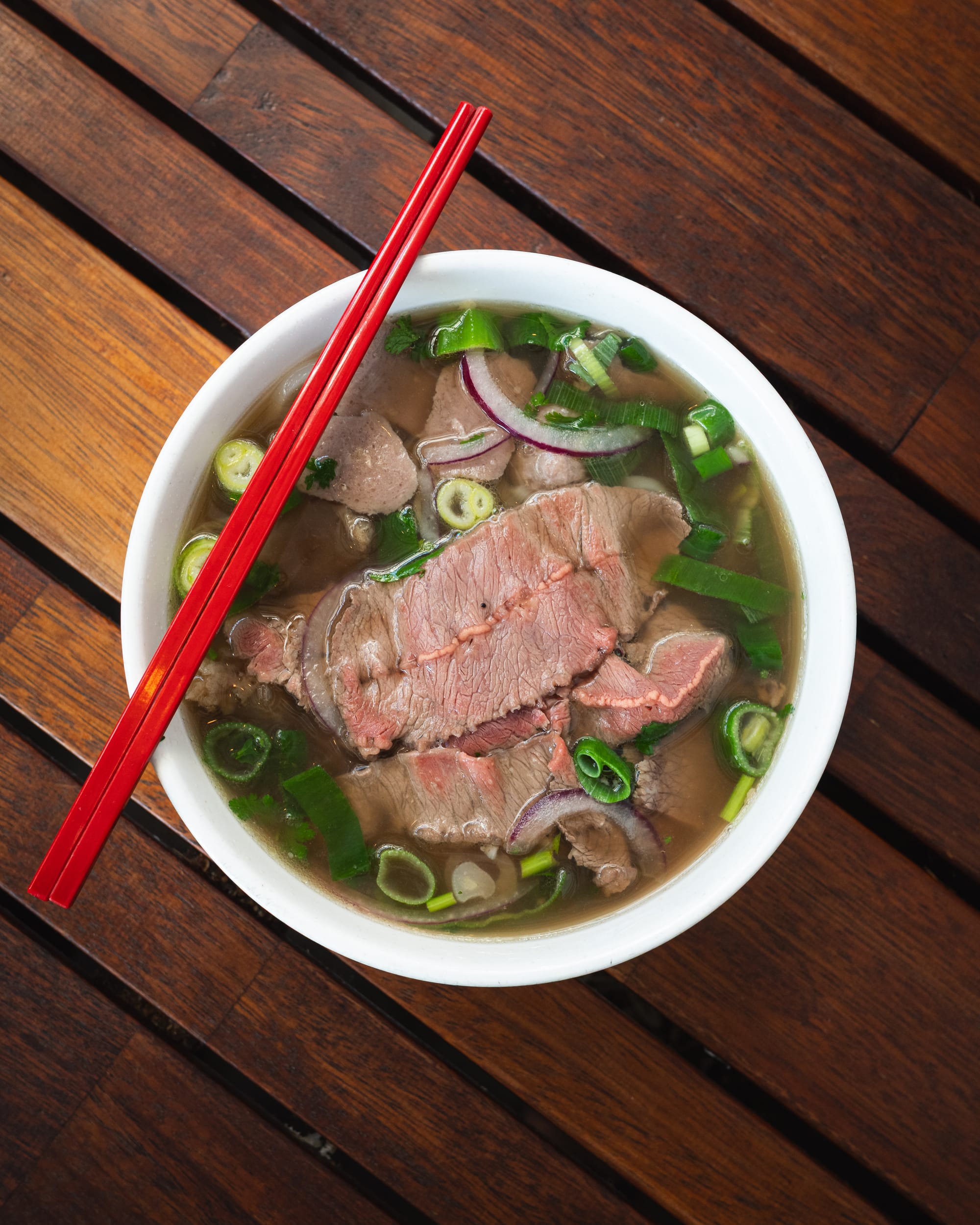 A bowl of Vietnamese pho with thinly sliced beef, green onions, and red chopsticks resting on the side.