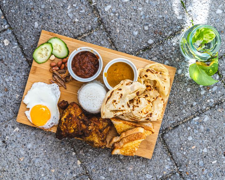 Chopping board on a brick floor with a plant to the right, filled with egg, fried chicken, toast, roti, rice and condiments