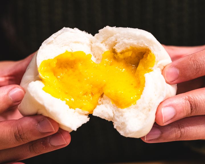 Hand holding a salted egg custard bun split open showing the oozing salted egg custard